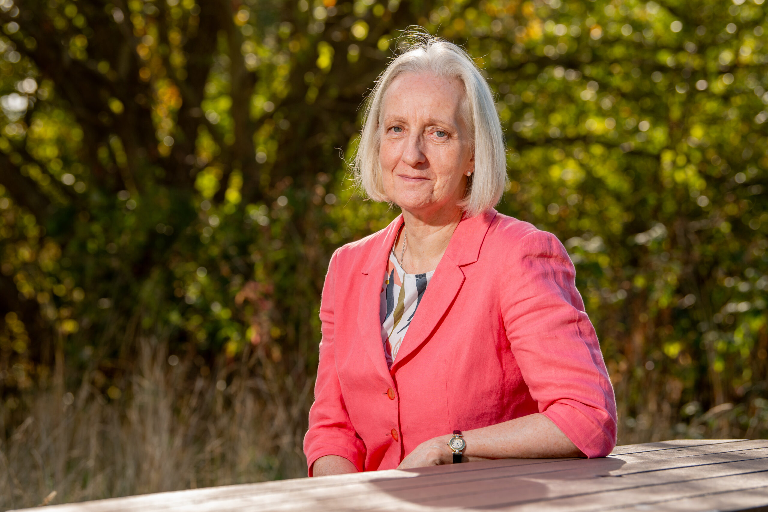 Anita Meston sitting at a wooden table outdoors, wearing a coral jacket